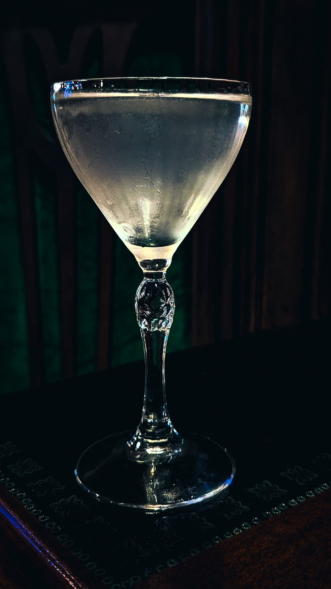 A frosted Nick and Nora glass with a clear cocktail against a dark green wall