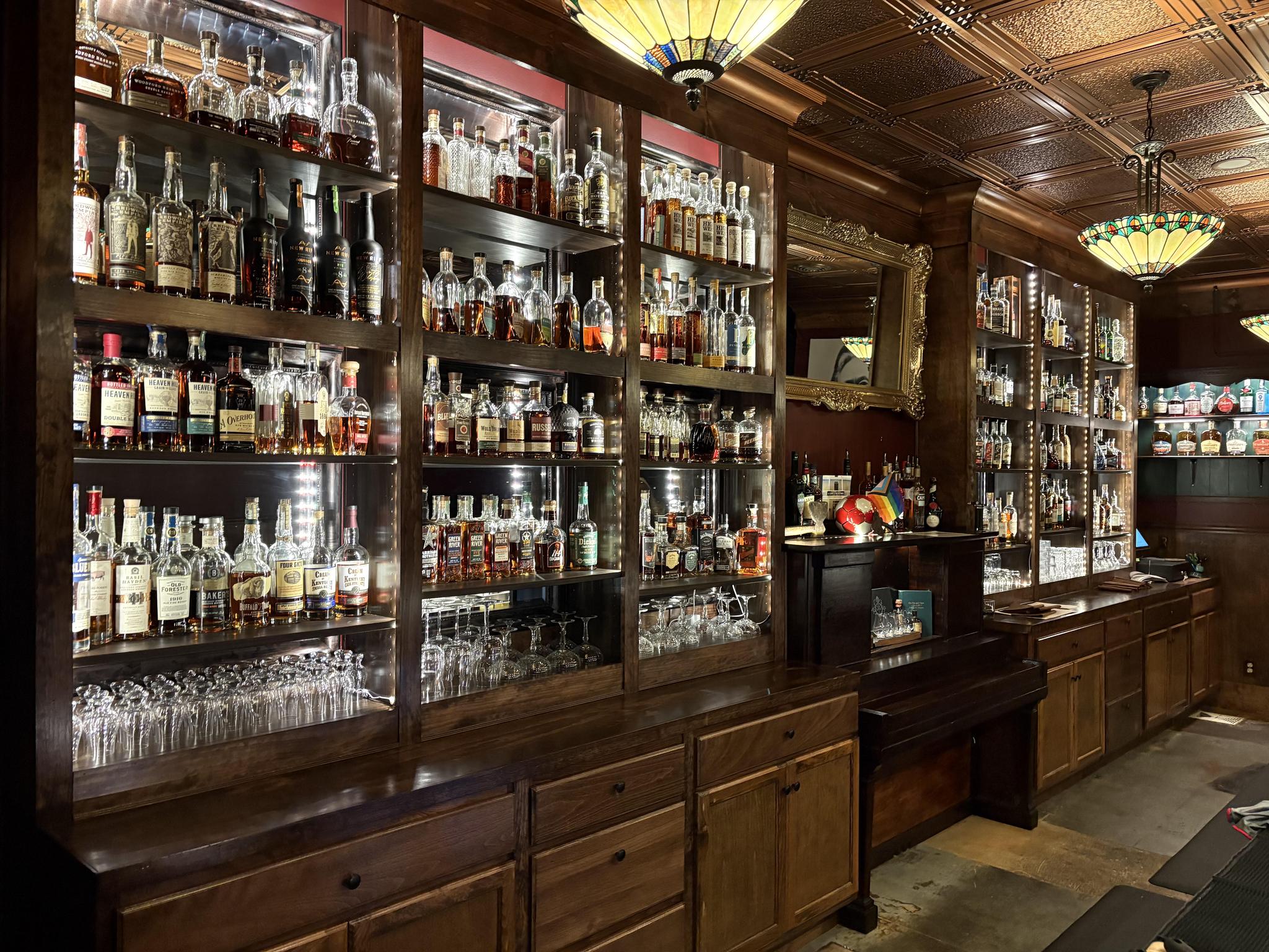 The back bar at Carrie’s: tall wooden shelves of lit whiskey bottles, rows of glassware below, a gilt mirror and stained-glass pendant lamps under a pressed-tin ceiling