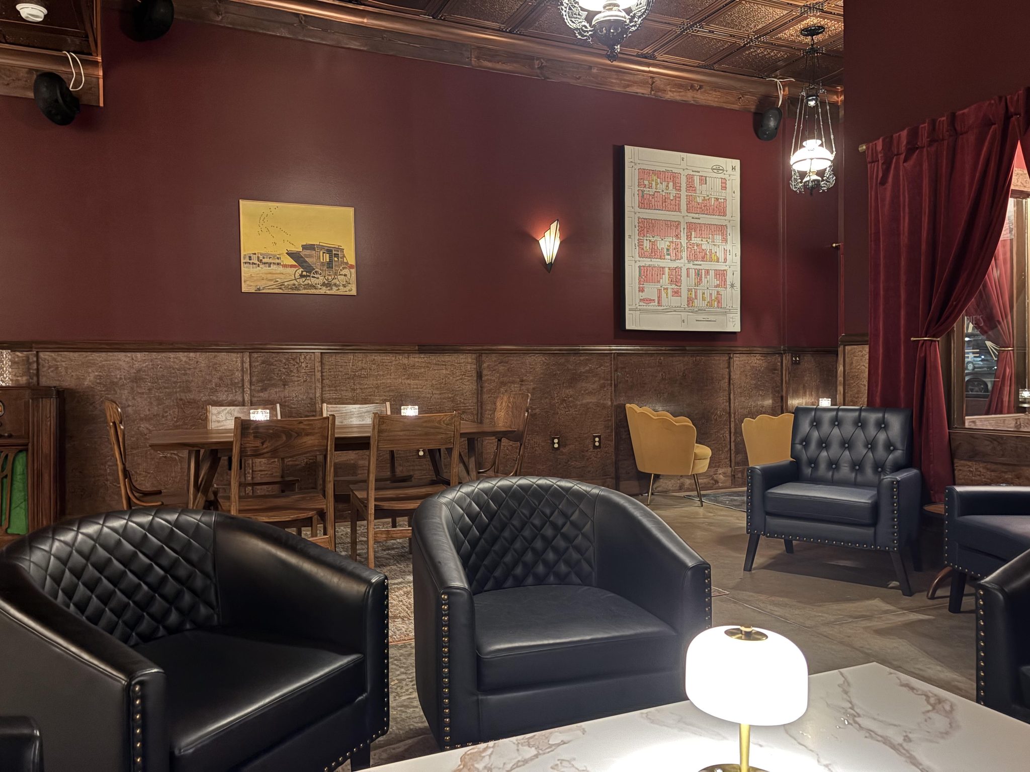 The lounge at Carrie’s: quilted leather club chairs around a marble table with a small lamp, a long wooden table with chairs along the wainscoted wall, a Deco sconce and a framed old street map on the red wall